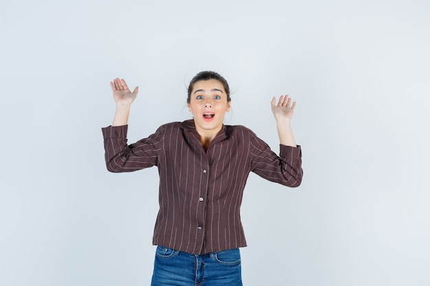 Young woman raising palms in surrender gesture in striped shirt, jeans and looking surprised , front view.