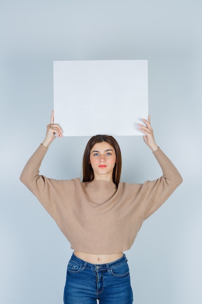 Foto gratuita giovane donna alzando il cartone in maglione, jeans e guardando malinconico, vista frontale.