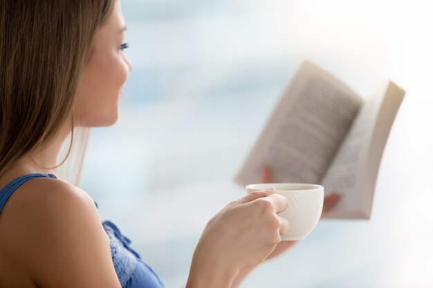 Young woman reading book, holding cup of coffee