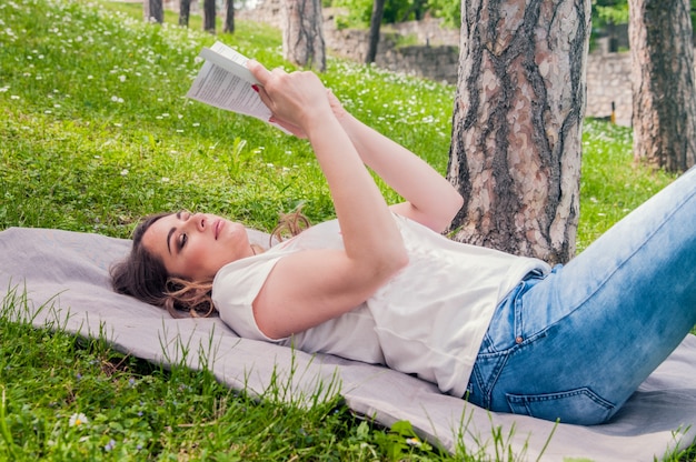 Молодая женщина, чтение книги в парке, лежа на траве. Селективный фокус. Молодая красивая внимательная женщина лежит на зеленой траве и читает книгу против городского парка.