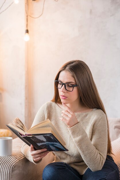 소파에 앉아있는 동안 책을 심각하게 읽는 젊은 여자