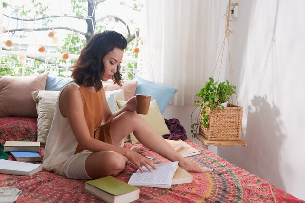 Young woman reading a book sitting on her bed with a cup of a drink