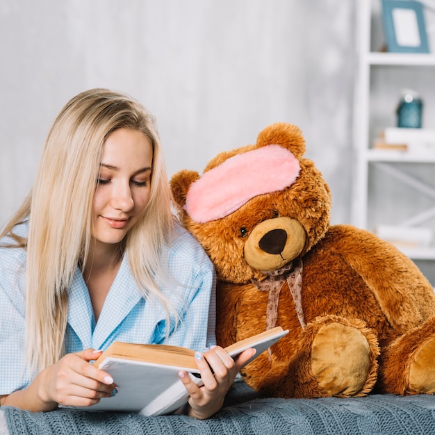 Young woman reading book with soft toy on bed