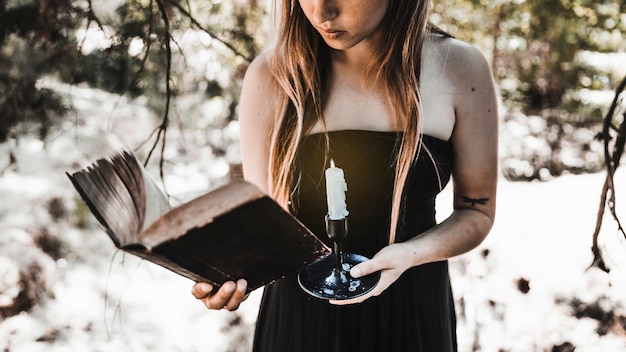 Young woman reading old book with candle in forest