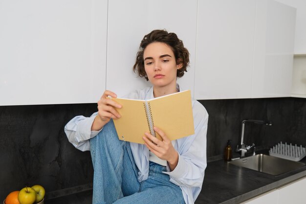 Молодая женщина с серьезным лицом читает свои заметки, сидит на кухонном столе и смотрит на готовящуюся тетрадь