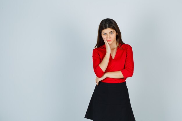 Young woman in red blouse, black skirt holding hand on cheek