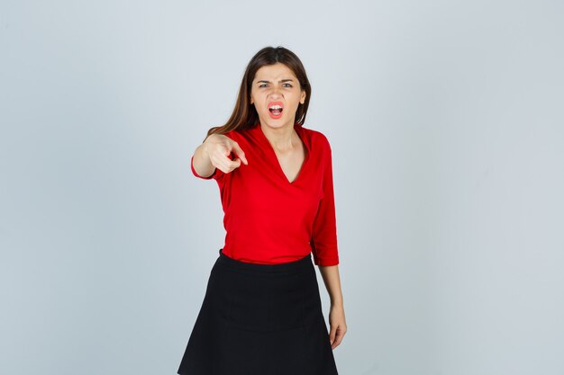 Young woman in red blouse, black skirt pointing at camera with index finger