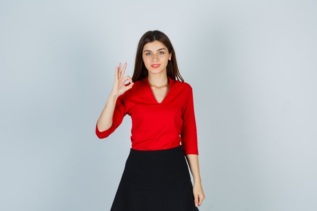 Young woman in red blouse, black skirt showing ok sign and looking happy