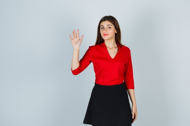 Young woman in red blouse, black skirt waving hand to greet and looking cute