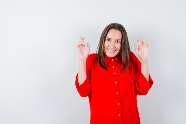 Young woman in red blouse keeping fingers crossed and looking merry , front view.