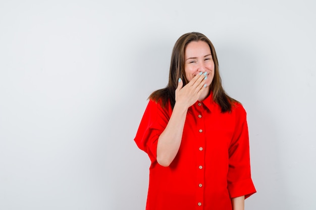 Young woman in red blouse keeping hand on mouth and looking merry , front view.