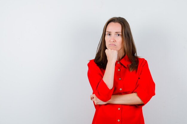 Young woman in red blouse leaning chin on fist and looking puzzled , front view.