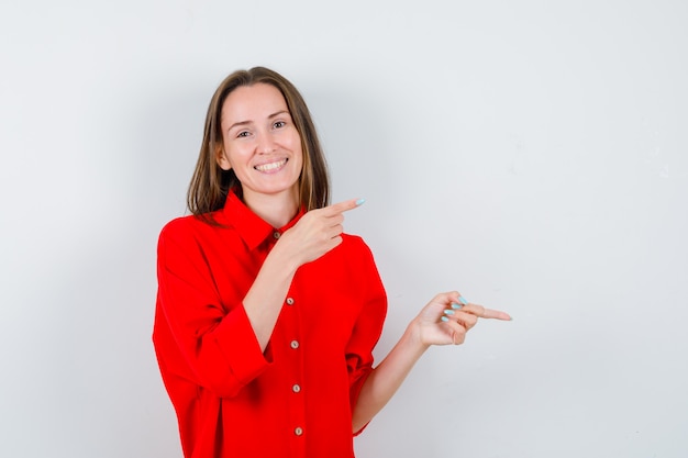 Young woman in red blouse pointing right and looking happy , front view.