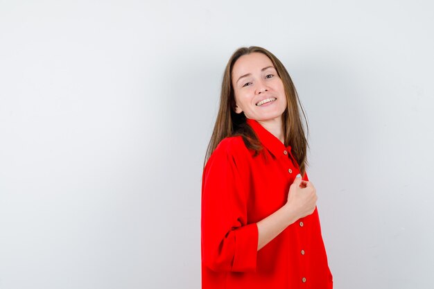 Young woman in red blouse posing, standing sideways and looking pleased .