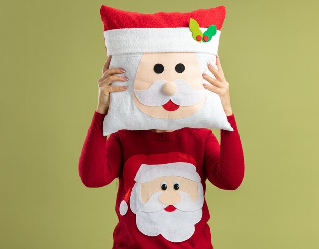 Free photo young woman in  red christmas sweater hiding her face with christmas pillow  standing over green wall