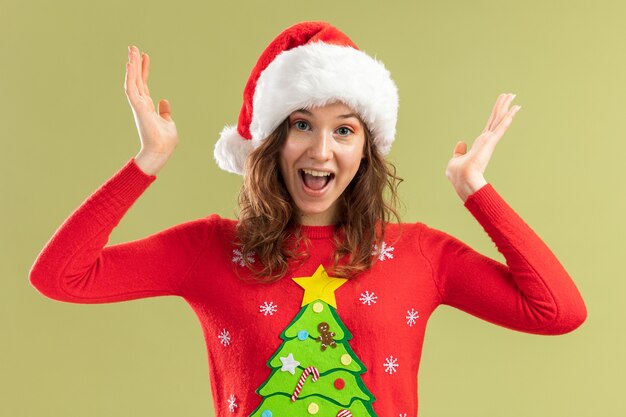 Молодая женщина в красном рождественском свитере и шляпе санта-клауса счастливая и взволнованная, поднимая руки, стоя на зеленом фоне