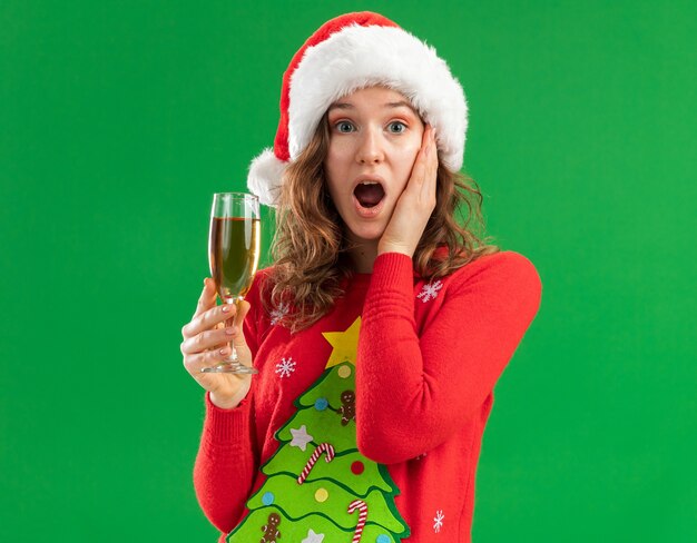 молодая женщина в красном рождественском свитере и шляпе санта-клауса держит бокал шампанского, глядя в камеру, изумленно положив руку на щеку, стоя на зеленом фоне