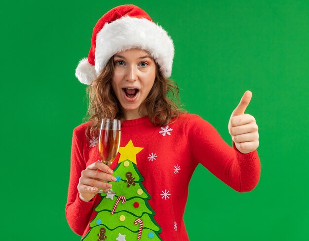 Молодая женщина в красном рождественском свитере и шляпе санта-клауса держит бокал шампанского, глядя в камеру, весело улыбаясь, показывая пальцы вверх, стоя на зеленом фоне