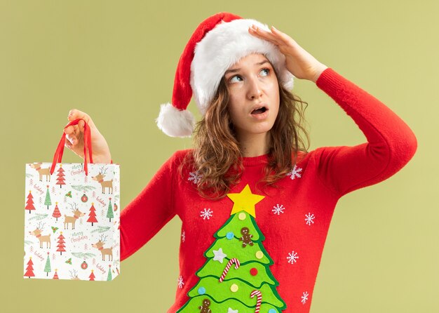 Молодая женщина в красном рождественском свитере и шляпе санта-клауса держит бумажный пакет с рождественскими подарками, озадаченно глядя вверх, стоя у зеленой стены