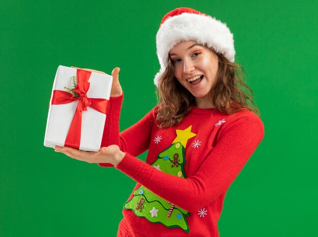 빨간 크리스마스 스웨터와 산타 모자에 젊은 여자 녹색 배경 위에 서 웃 고 행복 한 얼굴로 카메라를보고 선물을 들고