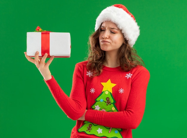 緑の背景の上に立っている懐疑的な表情でそれを見てプレゼントを保持している赤いクリスマスセーターとサンタ帽子の若い女性