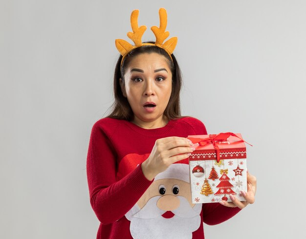 Young woman in red christmas sweater wearing funny rim with deer horns holding christmas gift looking being surprised 