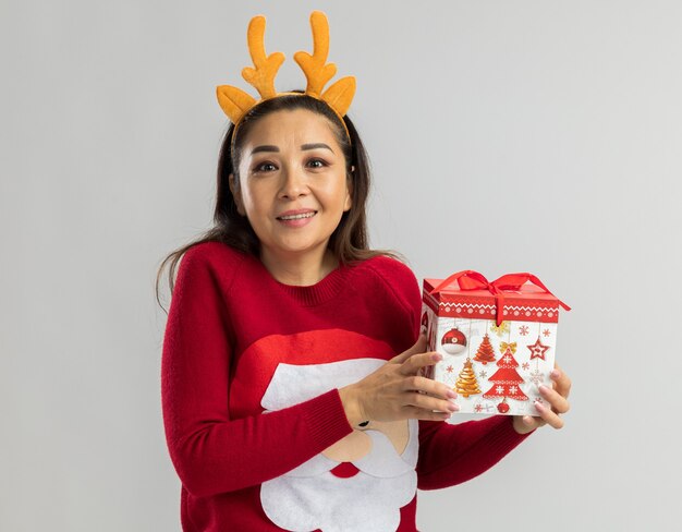 赤いクリスマスセーターの若い女性は、元気に幸せで前向きな笑顔に見えるクリスマスギフトを保持している鹿の角で面白いリムを身に着けています
