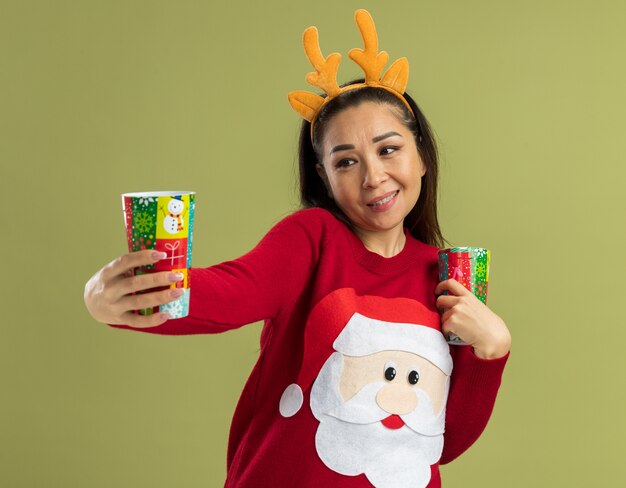 Молодая женщина в красном рождественском свитере в забавной оправе с оленьими рогами держит красочные бумажные стаканчики, счастливая и позитивная, весело улыбаясь
