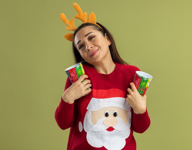 Young woman in red christmas sweater wearing funny rim with deer horns holding colorful paper cups looking feeling positive emotions smiling 