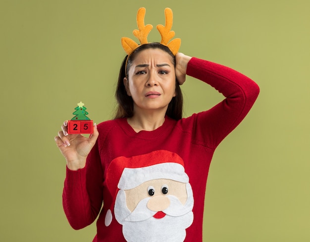 young woman in red christmas sweater wearing funny rim with deer horns holding toy cubes with date twenty five looking confused with hand on her head for mistake forgot standing over green background