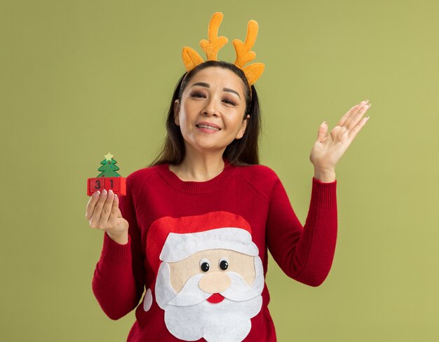 молодая женщина в красном рождественском свитере в забавной оправе с оленьими рогами держит игрушечные кубики с новогодней датой, глядя в камеру, счастливая и веселая улыбка с поднятой рукой, стоя на зеленом фоне