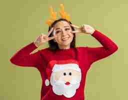 Free photo young woman in red christmas sweater wearing funny rim with deer horns looking at camera smiling with happy face showing v-sign near her eyes standing over green background