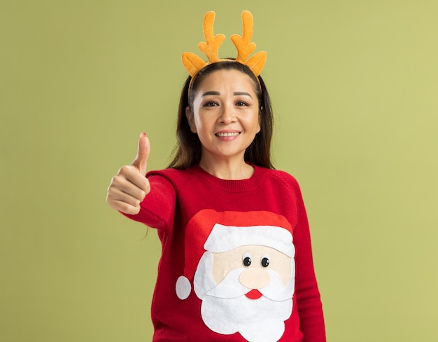 Young woman in red christmas sweater wearing funny rim with deer horns looking smiling showing thumbs up happy and positive 