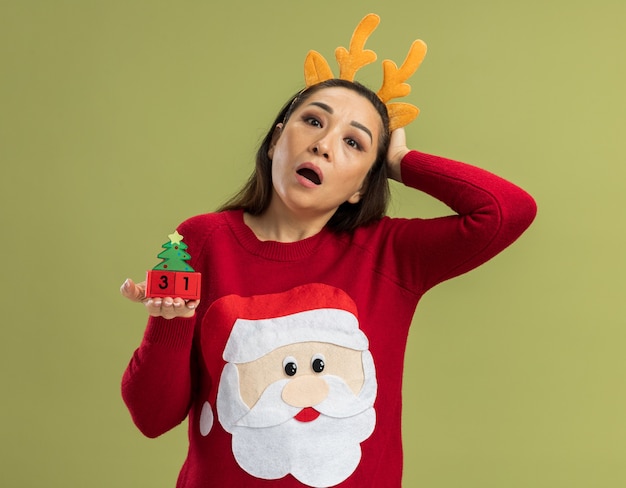 Молодая женщина в красном рождественском свитере в забавной оправе с оленьими рогами показывает игрушечные кубики с новогодней датой в изумлении, стоя у зеленой стены