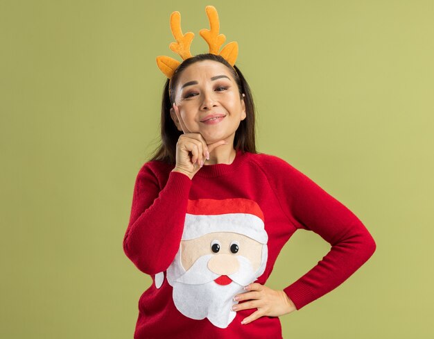 Young woman in  red christmas sweater wearing funny rim with deer horns smiling cheerfully happy and positive standing over green wall