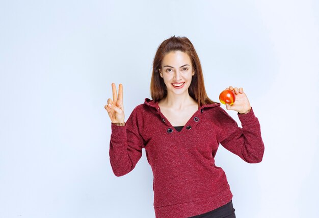 Young woman in red jacket holding a red apple and showing positive hand sign