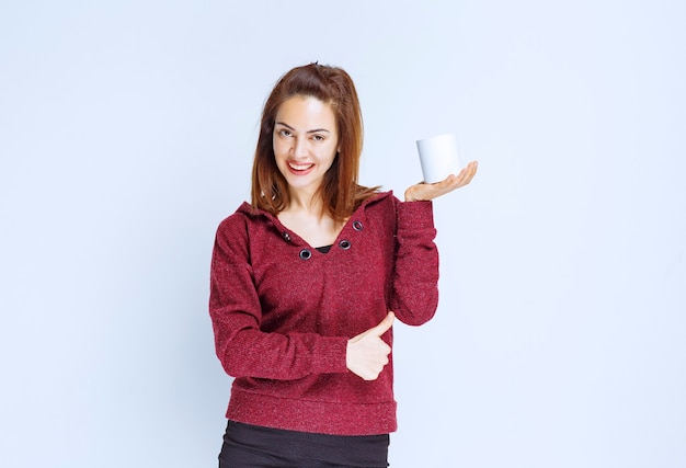 Young woman in red jacket holding a white coffee mug and showing positive hand sign