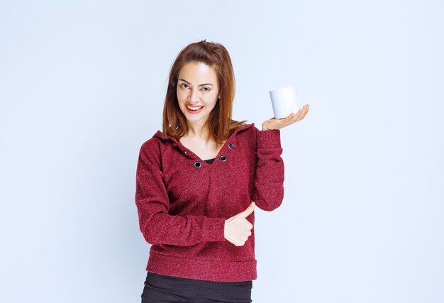 Молодая женщина в красной куртке держит кружку белого кофе и показывает знак рукой