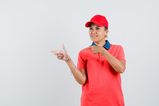 Young woman in red shirt and cap pointing left with index fingers and looking pretty , front view.