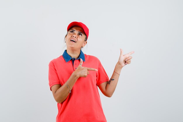 Young woman in red shirt and cap pointing right with index fingers and looking pensive , front view.