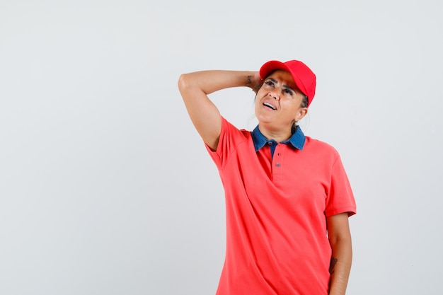 Young woman in red shirt and cap putting hand above head, thinking about something and looking pensive , front view.