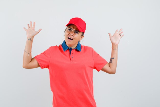 Young woman in red shirt and cap raising hands in surrender pose and looking pretty , front view.