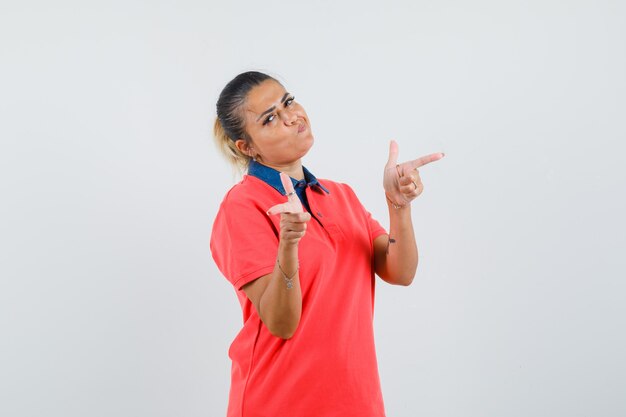 Young woman in red shirt pointing at different directions with index fingers and looking pretty , front view.