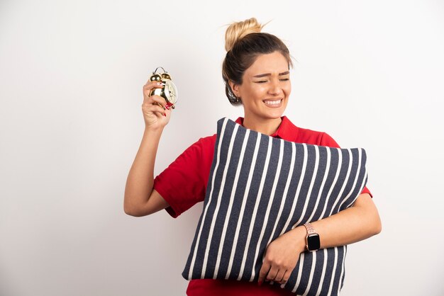 Young woman in red short showing an alarm clock and holding a pillow.