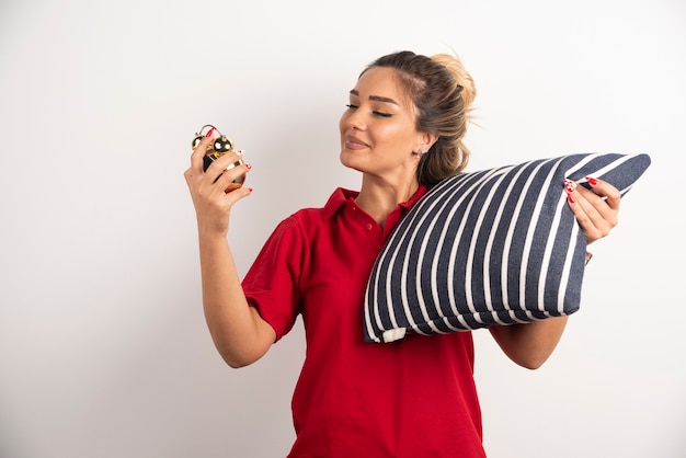 Молодая женщина в красном короткометражке показывает будильник и держит подушку.