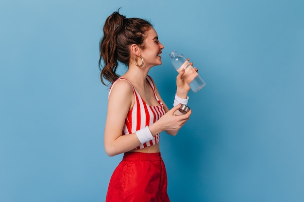 young woman in red striped outfit laughs and drinks water from bottle on isolated wall