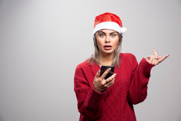 Молодая женщина в красном свитере держа мобильный телефон и смотря камеру.
