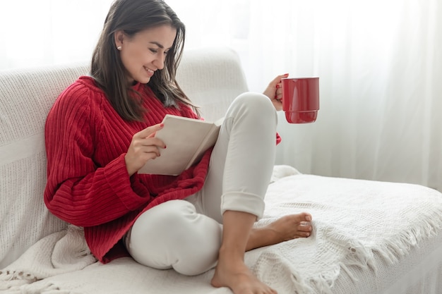 Young woman in a red sweater with a red cup reads a book.