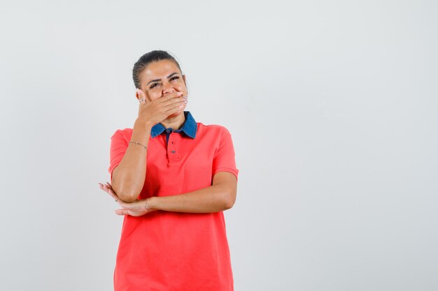 Young woman in red t-shirt covering mouth with hand and looking pretty , front view.