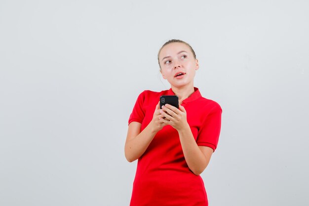 Молодая женщина в красной футболке держит мобильный телефон и выглядит задумчивой